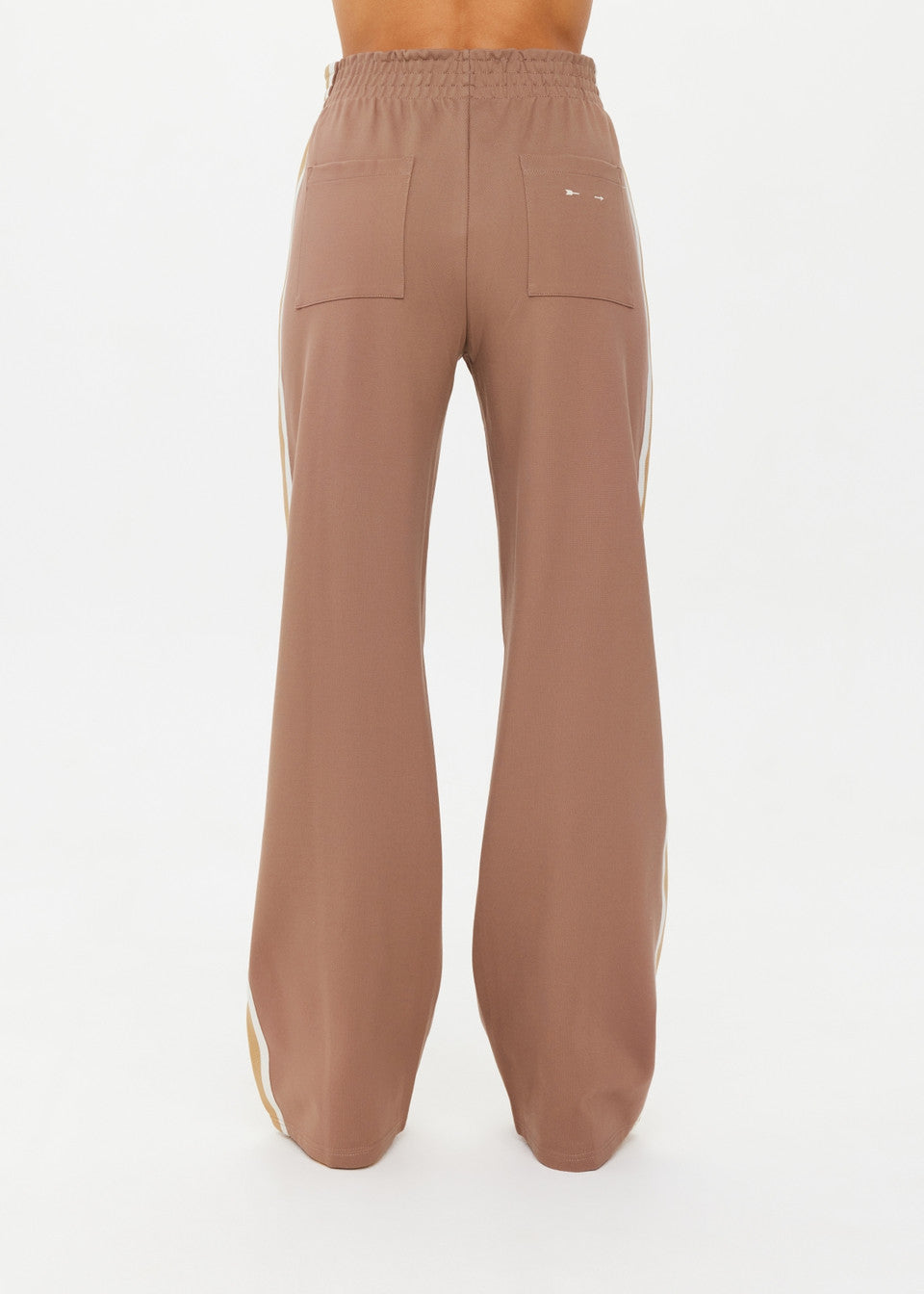 A person wearing The Upside's MONTE PANTS is shown from the back, against a white background. These high-waisted, loose-fitting pants are brown with wide legs and white side stripes, crafted from Lenzing viscose and featuring two back pockets.