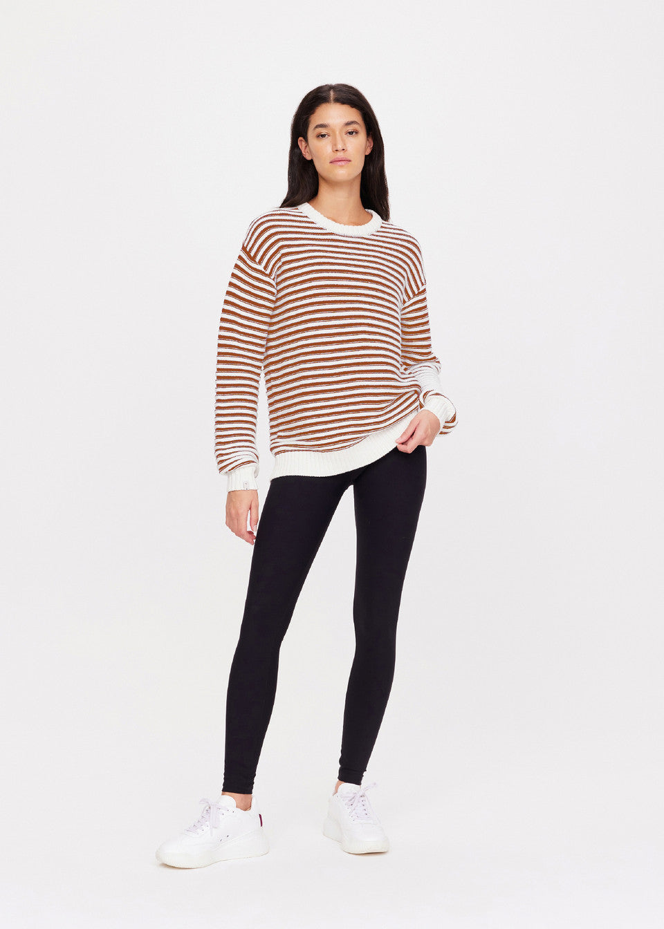 Against a white background, a person wears The Upside's BOO KNIT JUMPER in brown and white organic cotton stripes, paired with black leggings and white sneakers. Their long dark hair frames a neutral gaze towards the camera.