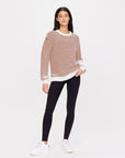 Against a white background, a person wears The Upside's BOO KNIT JUMPER in brown and white organic cotton stripes, paired with black leggings and white sneakers. Their long dark hair frames a neutral gaze towards the camera.