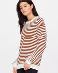 A person is wearing The Upside's BOO KNIT JUMPER, characterized by its classic brown and white horizontal stripes, stands against a plain white background, gazing slightly to the side.