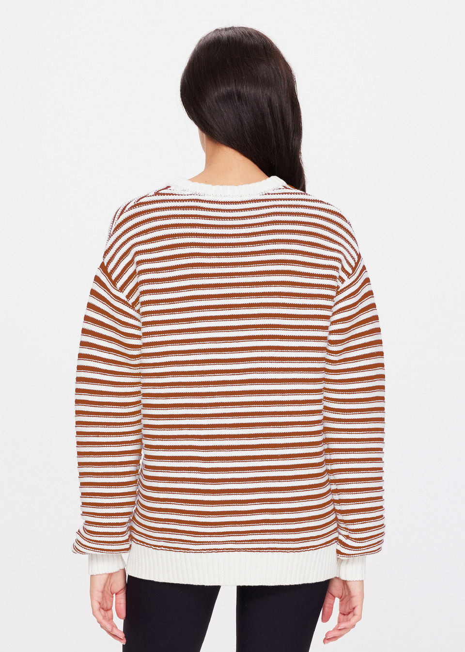 A person with long dark hair wears The Upside's BOO KNIT JUMPER, a white and brown striped sweater, paired with black pants. The plain white background accentuates the classic knitwear's timeless charm.