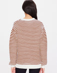 A person with long dark hair wears The Upside's BOO KNIT JUMPER, a white and brown striped sweater, paired with black pants. The plain white background accentuates the classic knitwear's timeless charm.