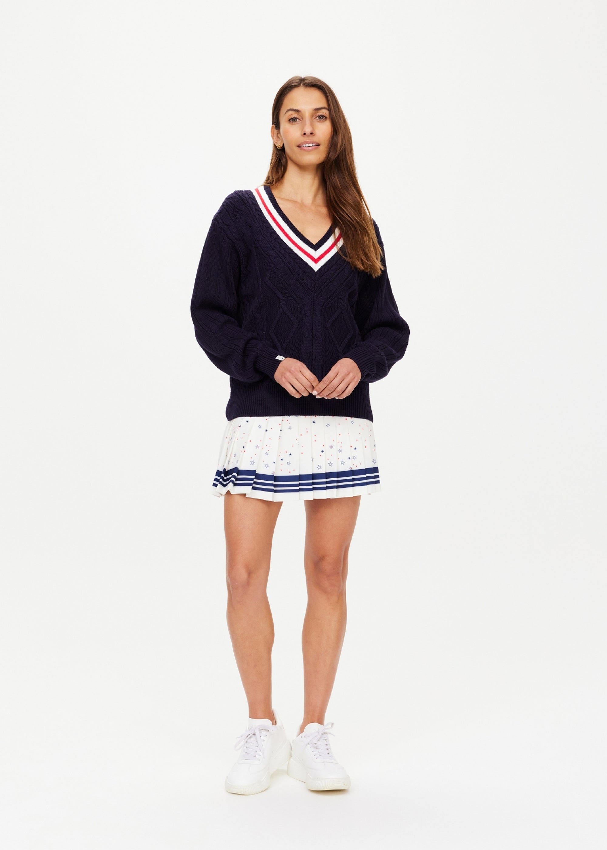 A woman with long brown hair wears The Upside's RENSHAW JOSETTE V NECK SWEATER, a navy knit with red and white trim, paired with a white pleated skirt featuring blue and red dots and white sneakers, standing against a plain white background.