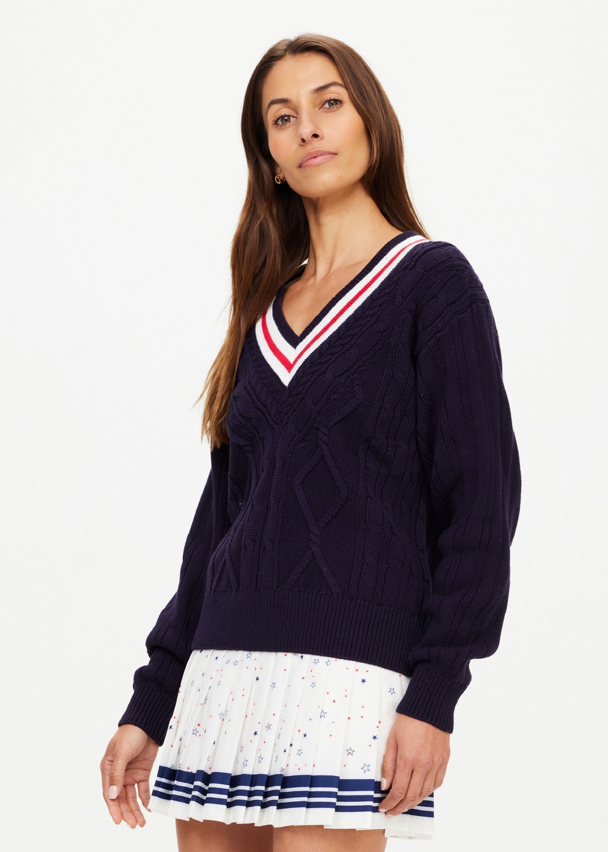 A woman with long brown hair models The Upside's RENSHAW JOSETTE V NECK SWEATER in navy blue, featuring intricate cable detailing, paired with a white pleated skirt with colorful prints against a plain light background.