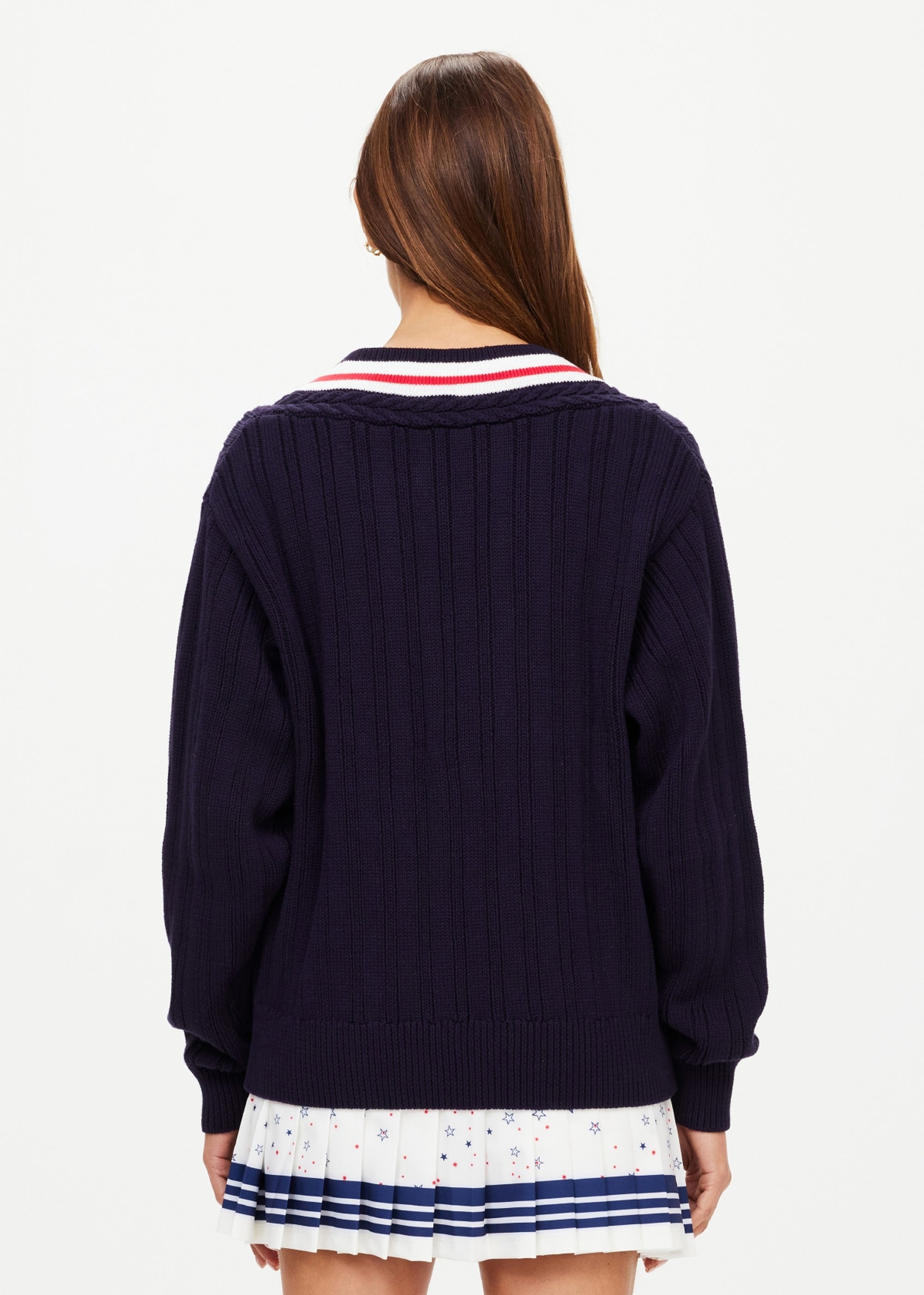 A woman with long brown hair faces away, wearing The Upside's RENSHAW JOSETTE V NECK SWEATER in navy with white, red, and navy collar detail, paired with a white pleated skirt featuring small colorful patterns.