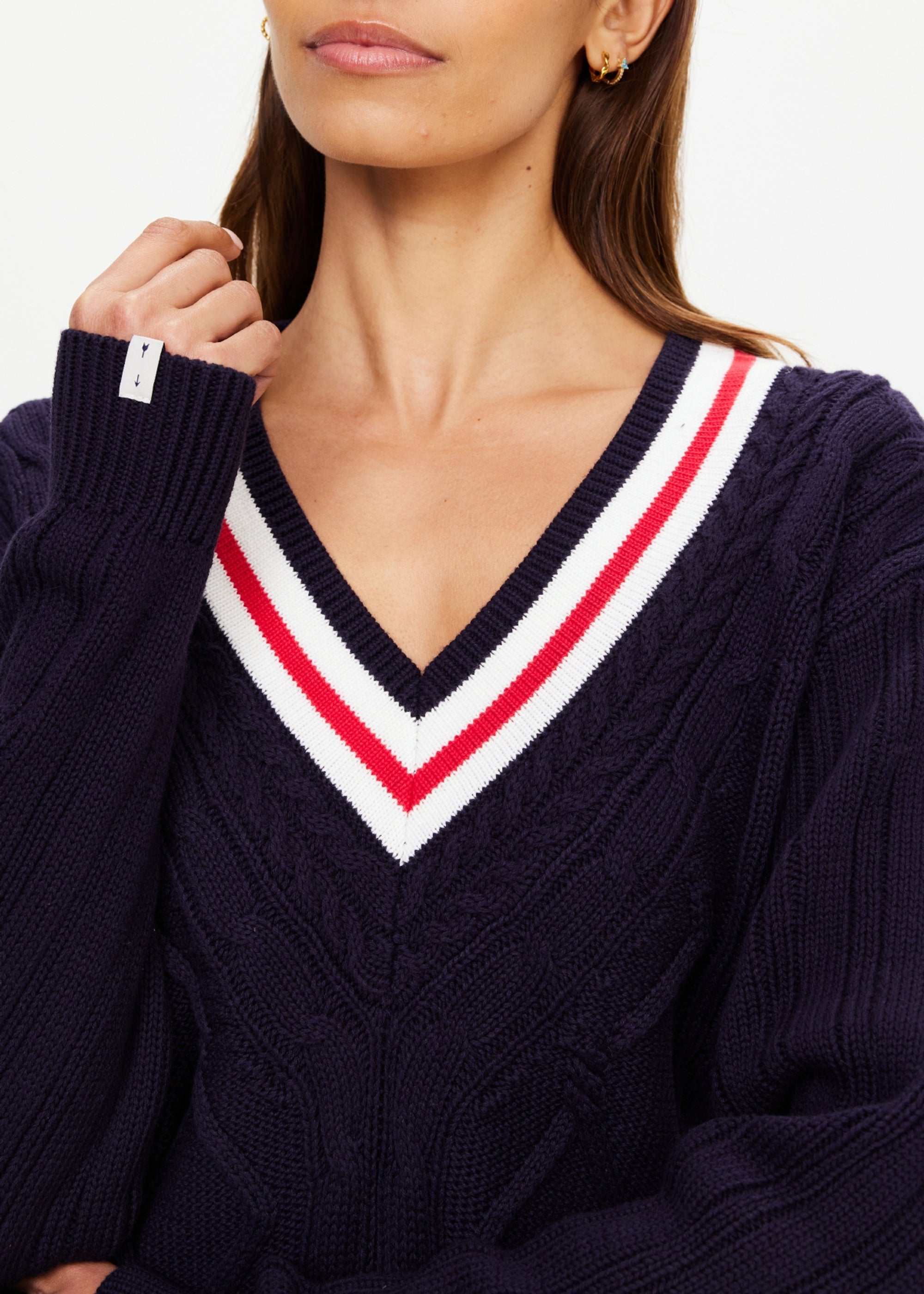 A woman wears The Upside RENSHAW JOSETTE V NECK SWEATER, an organic cotton navy blue piece with cable details and a deep V-neck accented by white and red stripes. She has light skin, long brown hair, and small gold hoop earrings.
