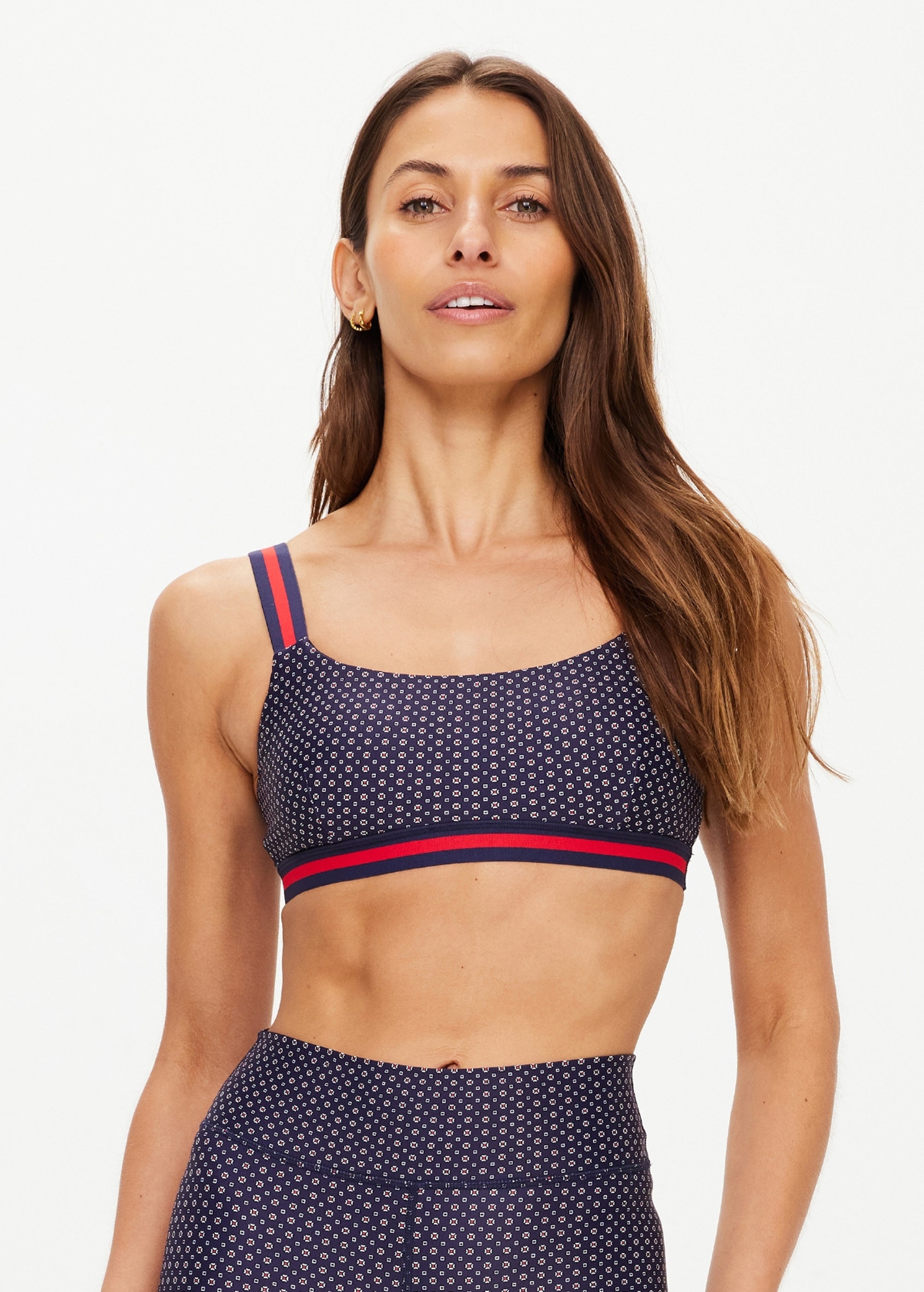 A woman with long brown hair wears The Upside's CAVALLO SCOOP NECK NINA BRA—a navy geometric print, quick-drying, mid coverage sports bra with red and navy striped straps—plus matching leggings, against a light background. Stylish and functional.