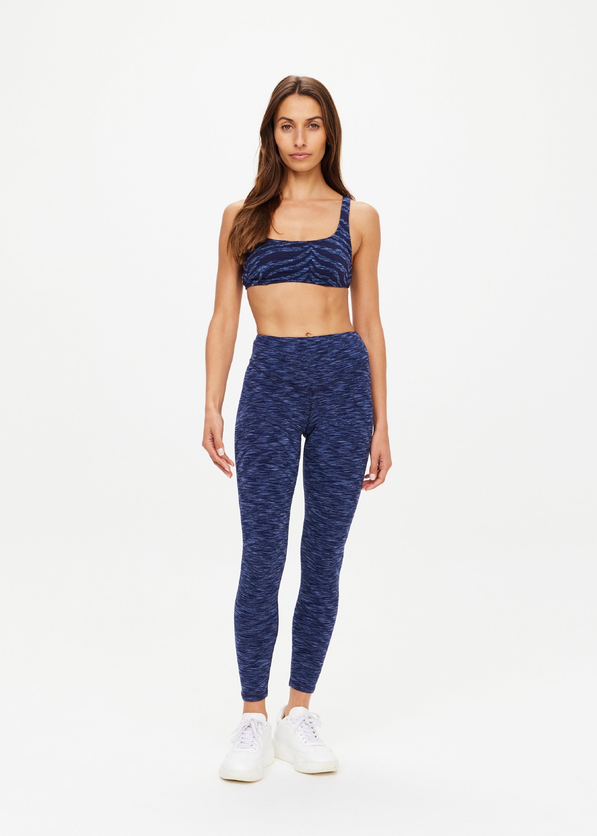 A woman faces forward in The Upside’s MYSTERE REVERSIBLE RORY BRA and matching high-waisted, space dye blue leggings with a subtle pattern and moisture-wicking fabric, styled with white sneakers against a light backdrop.