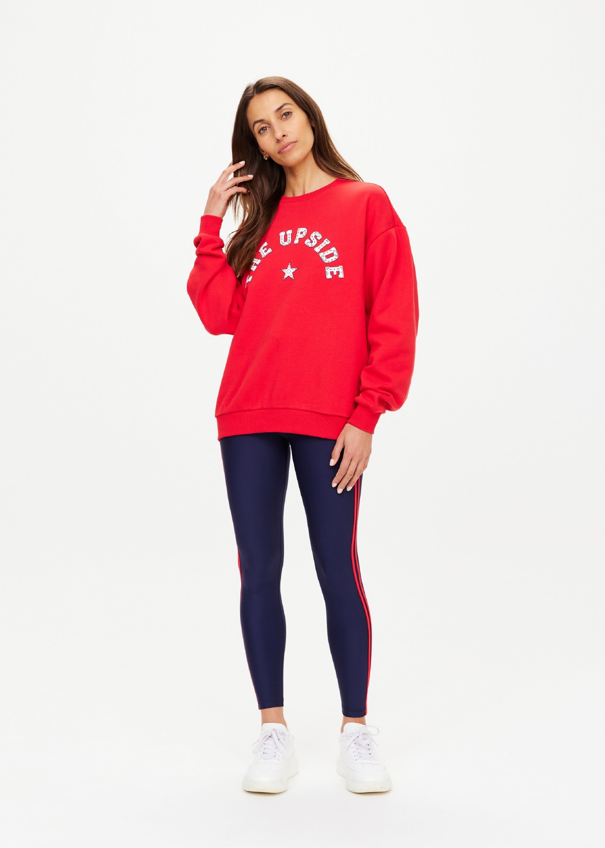 A woman with long brown hair poses against a plain background in The Upside’s RANCHERO SATURN CREW SWEATER, featuring organic cotton and the Western Logo, styled with navy leggings with red stripes and white sneakers.