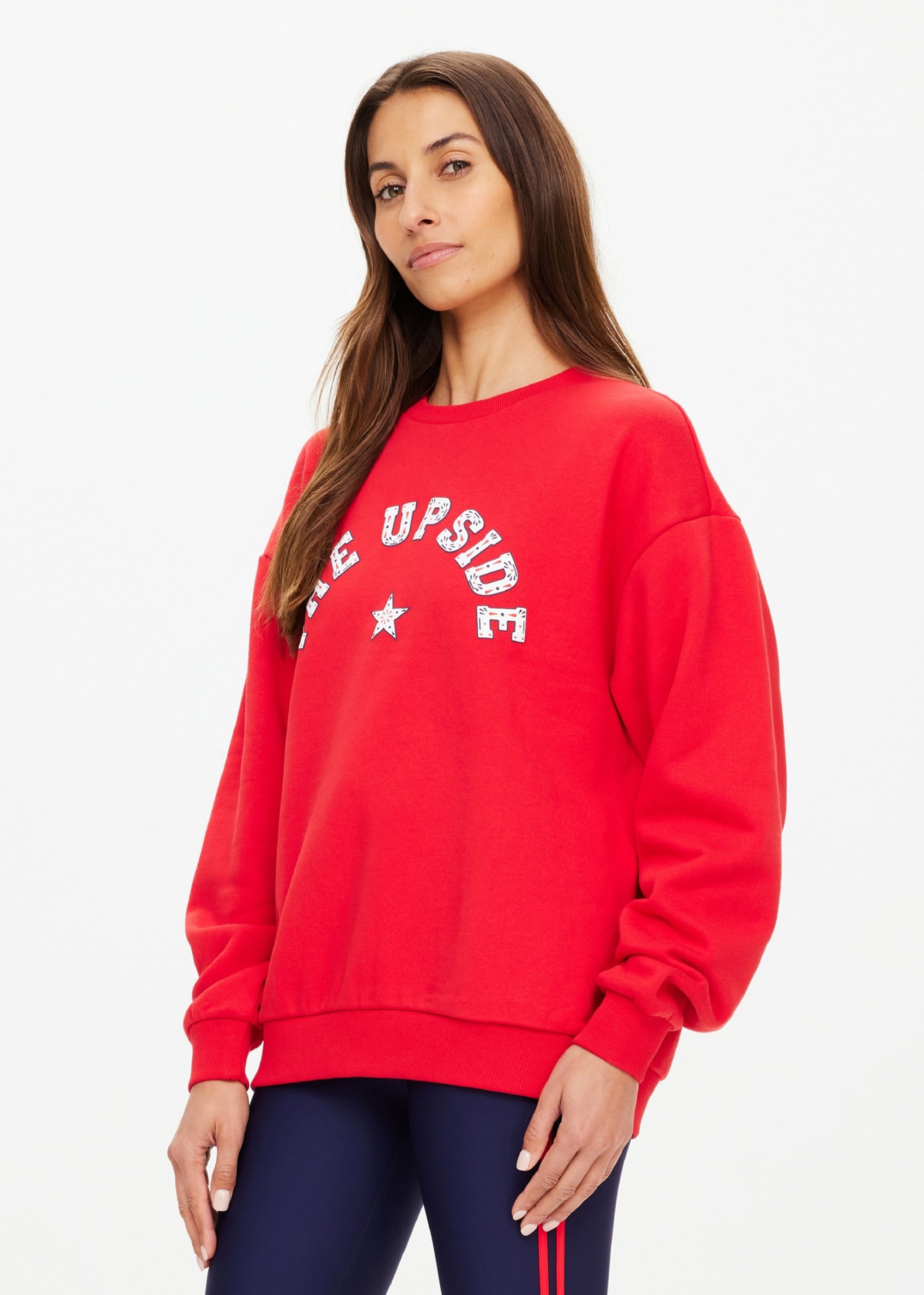 A woman with long brown hair wears The Upside's RANCHERO SATURN CREW SWEATER, a relaxed red organic cotton sweatshirt featuring a Western logo and star, paired with dark leggings, standing against a plain white background.