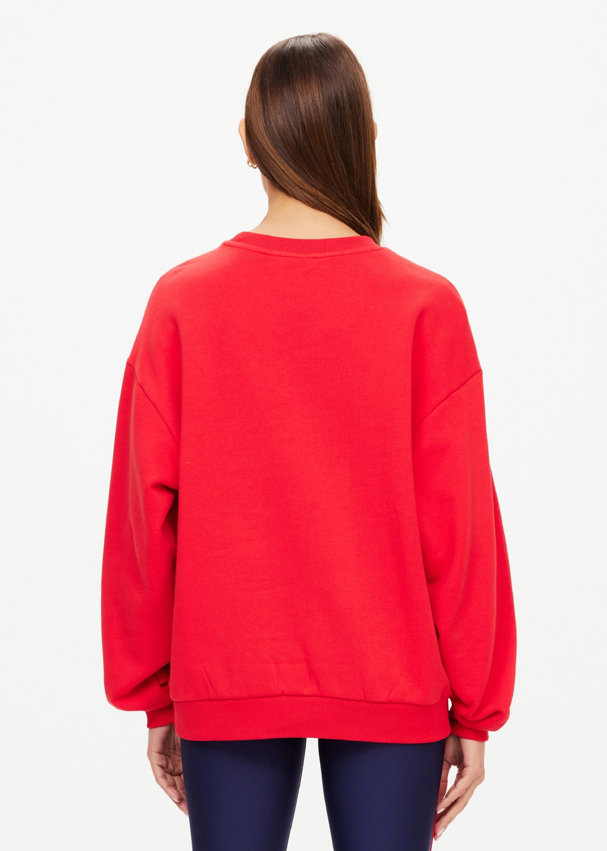A person with straight brown hair wears The Upside's RANCHERO SATURN CREW SWEATER in bright red organic cotton, featuring a subtle puff print logo, paired with dark blue pants. They stand back to the camera against a light background.