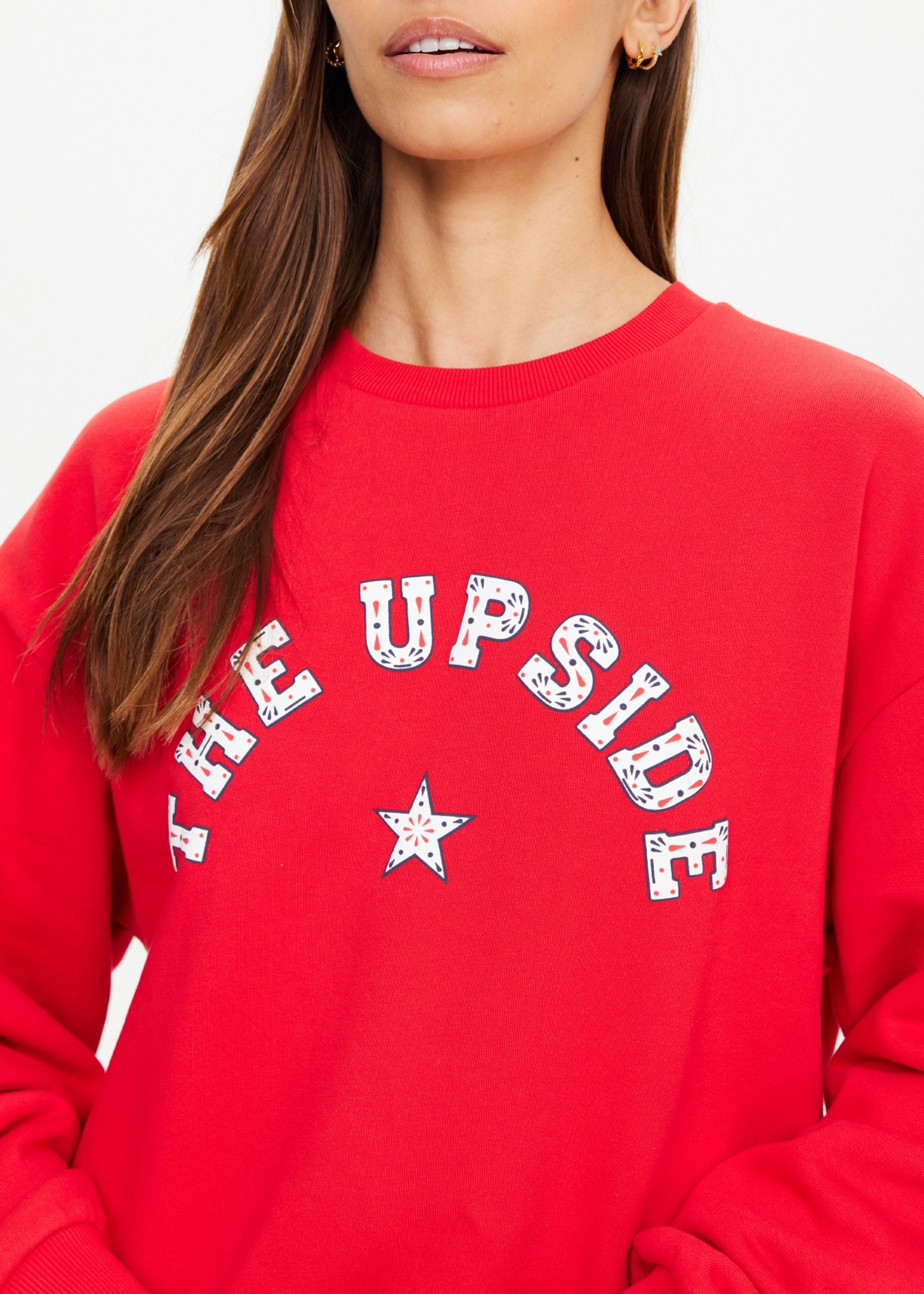 A woman wears The Upside's RANCHERO SATURN CREW SWEATER, a bright red organic cotton sweatshirt featuring a Western-style "THE UPSIDE" and star logo across the front. Only her lower face and upper torso show against a plain background.