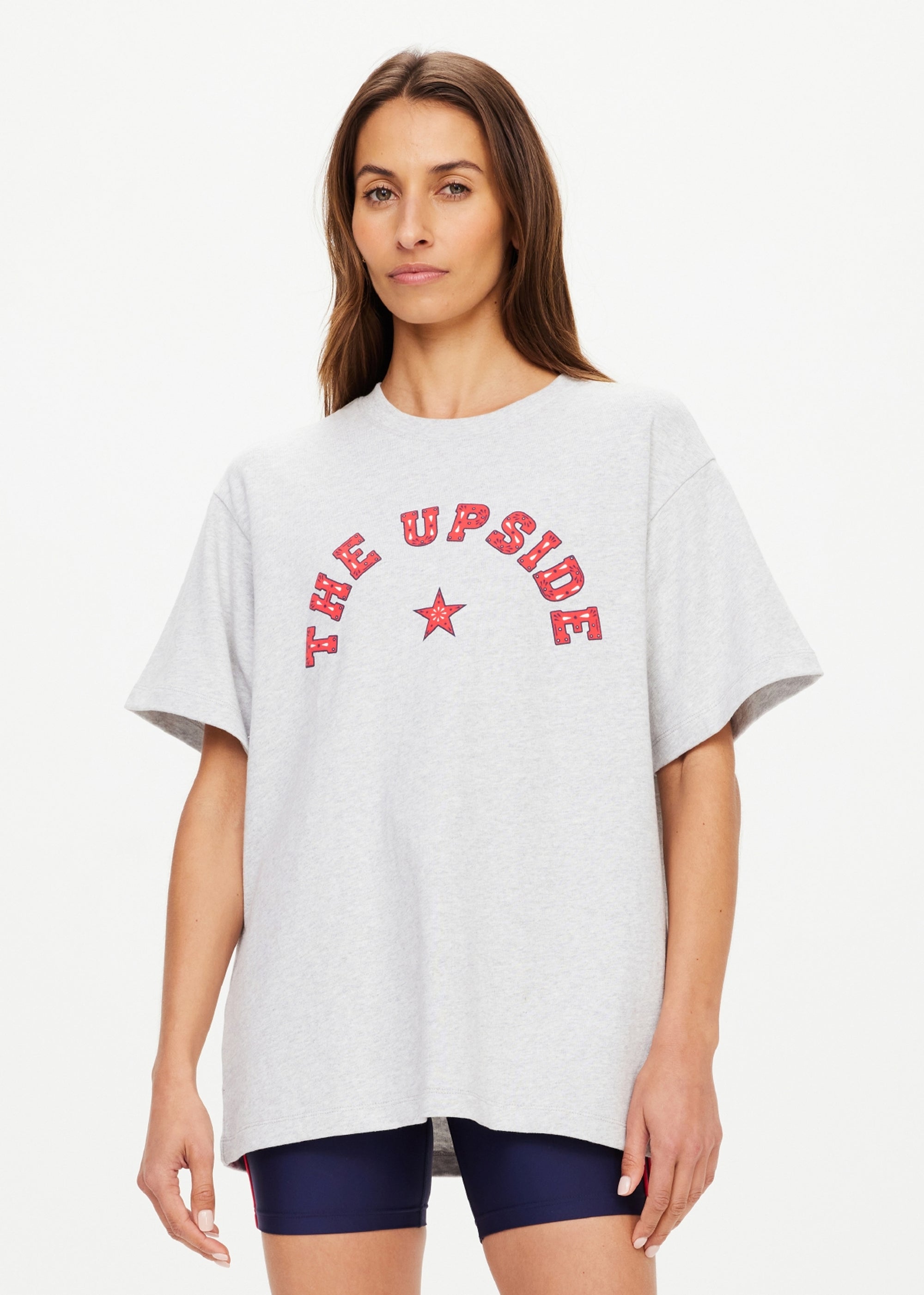 A woman with long brown hair wears The Upside's RANCHERO SAMMI TEE, an oversized organic cotton light gray top with a "THE UPSIDE" and red star Western logo, paired with dark shorts against a plain light background.