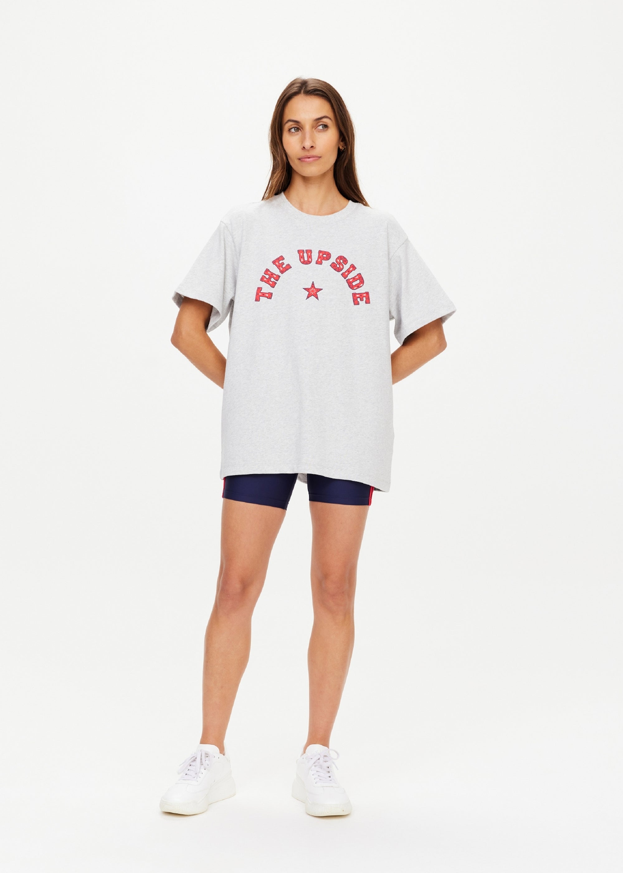 A woman stands against a plain background wearing The Upside’s RANCHERO SAMMI TEE—an oversized grey organic cotton T-shirt with a red "THE UPSIDE" Western logo and star—paired with navy shorts and white sneakers, hands behind her back.