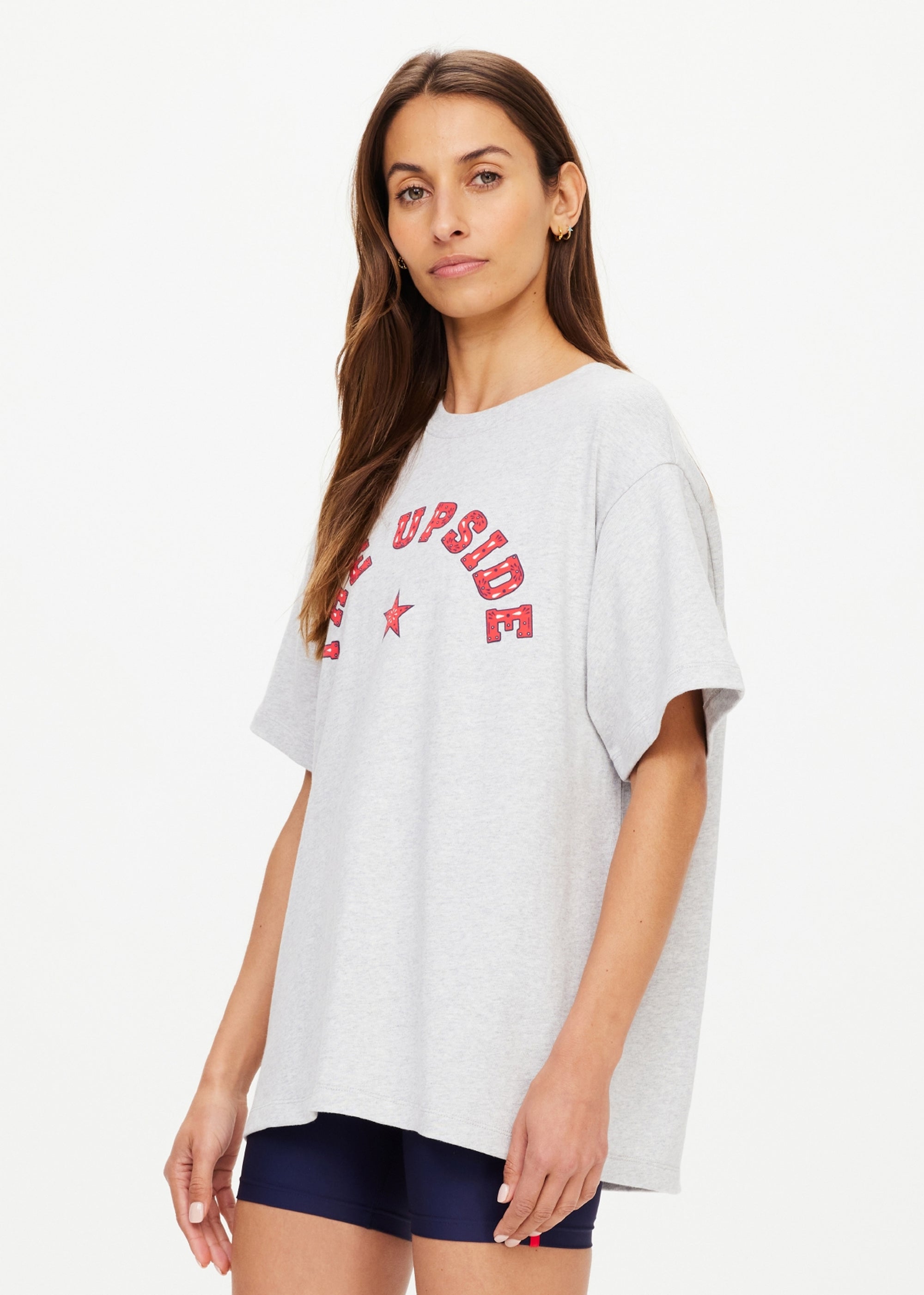 A woman with long brown hair wears The Upside’s RANCHERO SAMMI TEE, an oversized grey organic cotton T-shirt with a red western "THE UPSIDE" logo and star, paired with navy shorts, standing against a plain light background.
