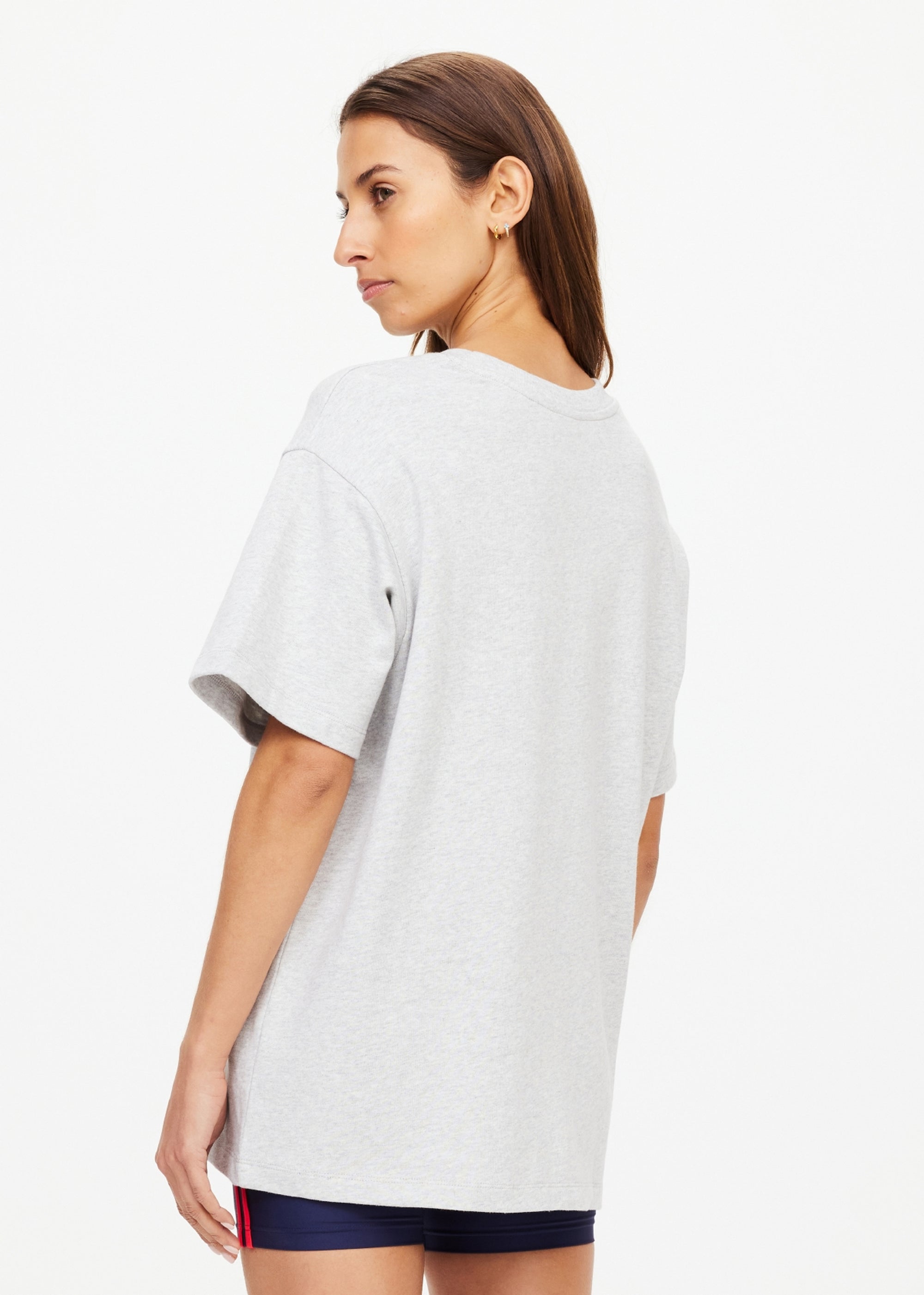 A woman with straight brown hair stands facing away, wearing The Upside RANCHERO SAMMI TEE in light gray organic cotton and navy shorts with red side detailing, against a plain white background.