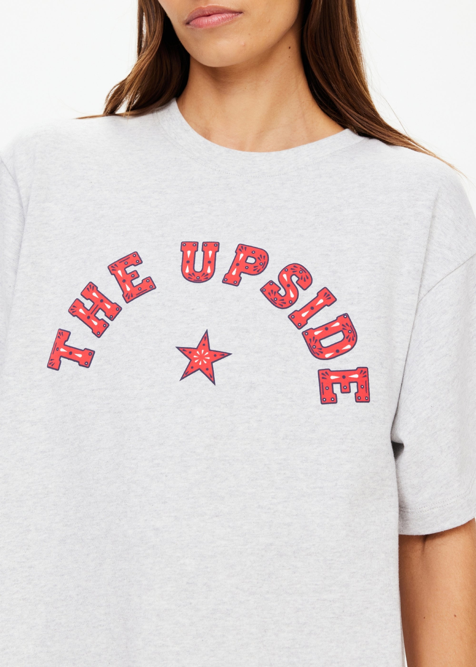 A person wearing The Upside's RANCHERO SAMMI TEE, an oversized organic cotton light grey top featuring a red star Western logo and "THE UPSIDE" on the front. Only the lower half of the face and upper torso are shown.