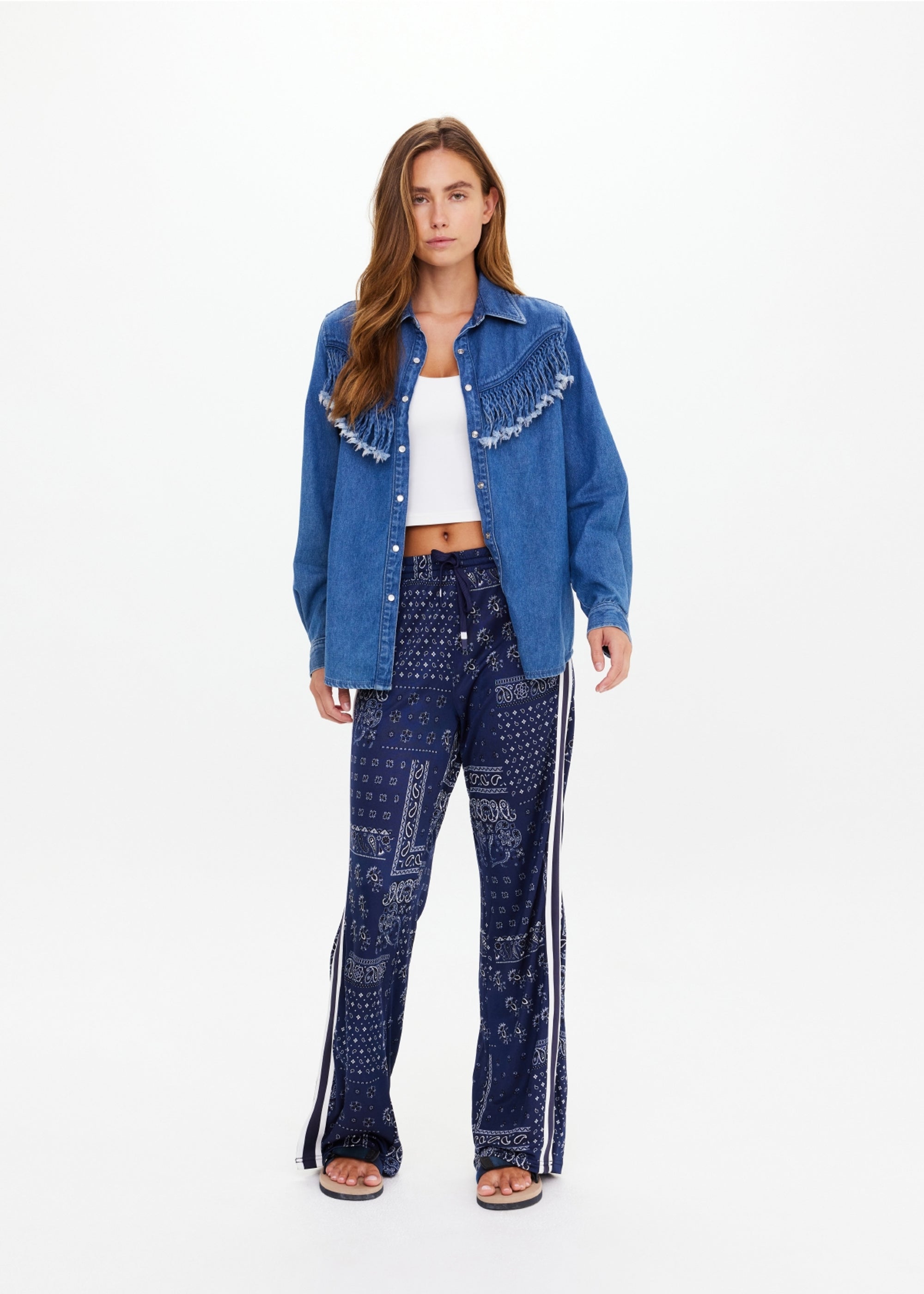 A woman faces forward against a plain white background, wearing The Upside WILDWEST SYLVE FRINGE SHIRT in blue chambray with fringe, a white crop top, patterned blue bell-bottom pants with white side stripes, and black sandals.