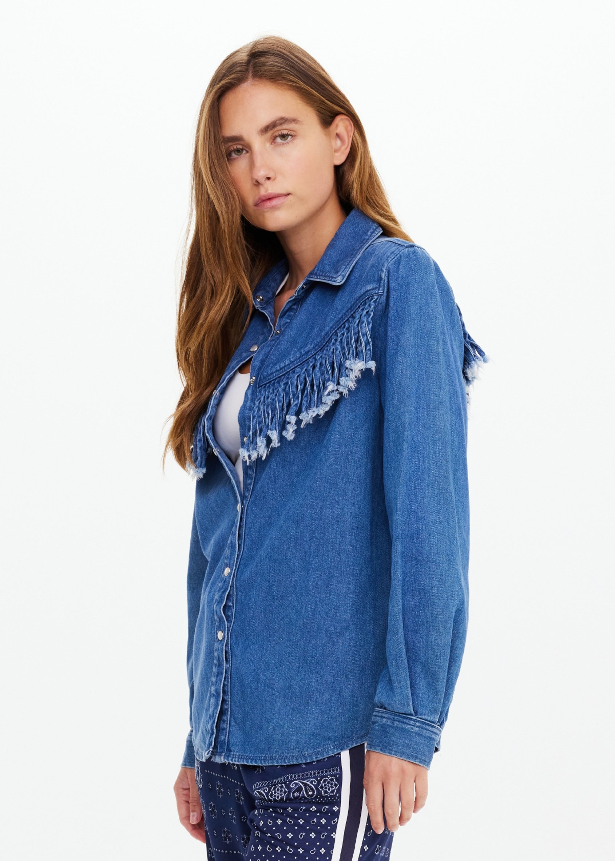 A woman with long brown hair models The Upside’s WILDWEST SYLVE FRINGE SHIRT, a western-inspired chambray top with fringe on the front and sleeves, paired with patterned navy pants. She stands slightly sideways against a plain light background.