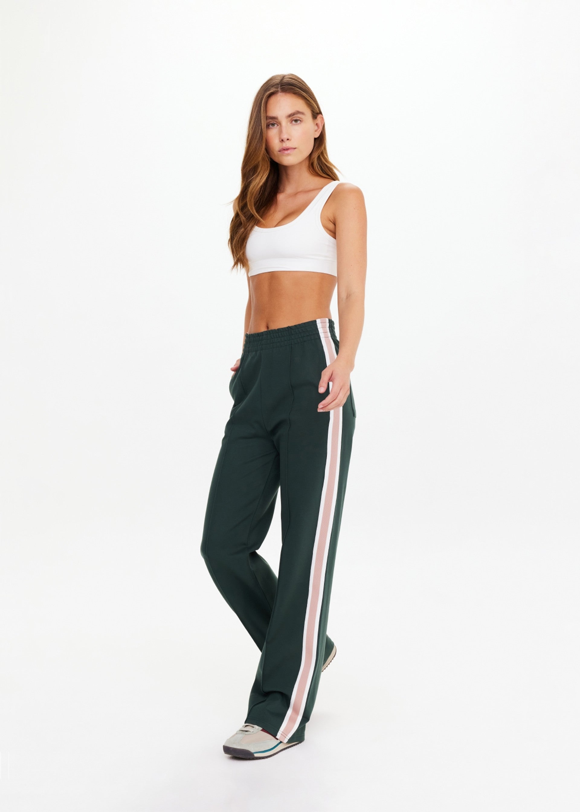 A woman wears The Upside MONTE PANT—high-waisted, wide leg with white and pink stripe tape—paired with a white sports bra and sneakers, standing with one hand in her pocket against a plain white background.