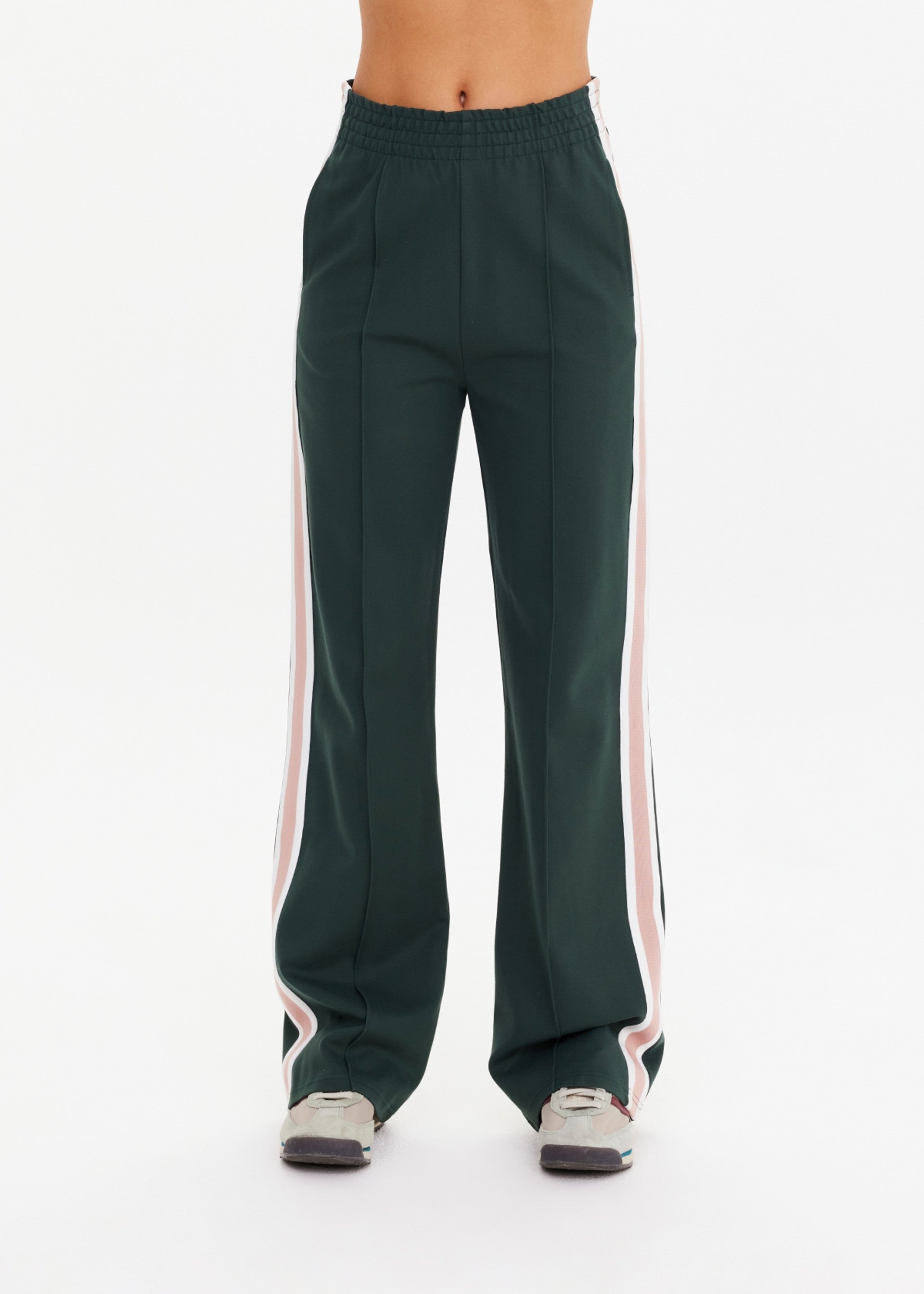 A person wearing The Upside MONTE PANT—high-waisted wide leg track pants with white and pink stripe tape—and gray sneakers, standing against a plain white background. Image is cropped above the waist.