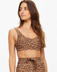 A woman with long brown hair wears The Upside’s Panthera Daisy Scoop Neck Bra in leopard print, made from recycled soft peached fabric, looking to her left against a plain white background.