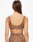 A woman with long brown hair is seen from the back, wearing The Upside Panthera Daisy Scoop Neck Bra in leopard print crafted from recycled soft peached fabric, paired with a high-waisted skirt against a plain white background.