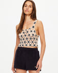 A woman with long brown hair wears The Upside's LUMINARY LILY CROCHET TOP, a sleeveless boho floral piece, paired with black shorts against a plain light background.