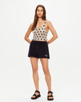 A woman stands against a plain white background wearing The Upside’s Luminary Lily Crochet Top, paired with black shorts and black sandals, her hands behind her back.
