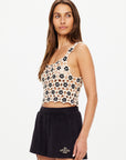 A woman with long brown hair wears The Upside's LUMINARY LILY CROCHET TOP and black shorts featuring the brand name, standing against a plain light background.