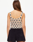 A woman, with straight brown hair, faces away from the camera wearing The Upside's LUMINARY LILY CROCHET TOP and high-waisted black shorts, creating a chic boho look against a plain white background.