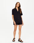 A woman with long, straight brown hair and a neutral expression models The Upside's SADDLE UP COLLARED HARDY SHIRT, a loose-fitting black button-up, paired with black shorts and sandals against a plain white background.