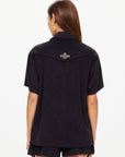 A person with long brown hair wears The Upside's SADDLE UP COLLARED HARDY SHIRT and matching shorts in black organic cotton. An embroidered white "THE UPSIDE SPORT" logo appears on the back as they face away from the camera.