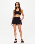 A woman stands against a plain white background wearing The Upside SADDLE UP ZIGGY SHORT, paired with a black sports bra and sandals, one hand in her pocket and a neutral expression.