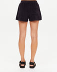 A person faces away from the camera, wearing The Upside SADDLE UP ZIGGY SHORT in black terry towelling with a small back pocket and black sandals, set against a plain white background.