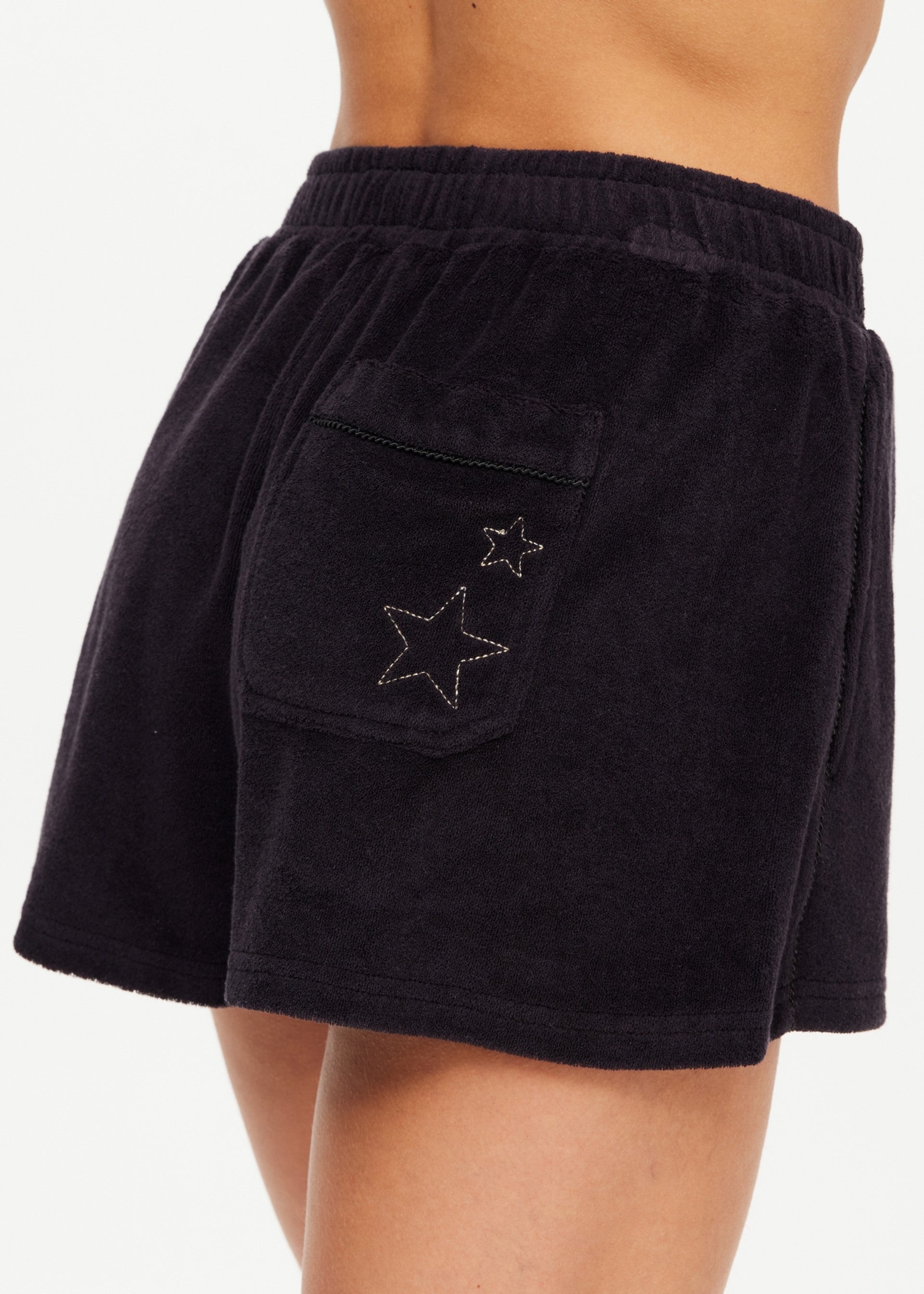 A close-up of a person wearing The Upside SADDLE UP ZIGGY SHORT—black terry towelling gym shorts featuring an elastic waistband and a back pocket with two star-shaped embroidered designs.