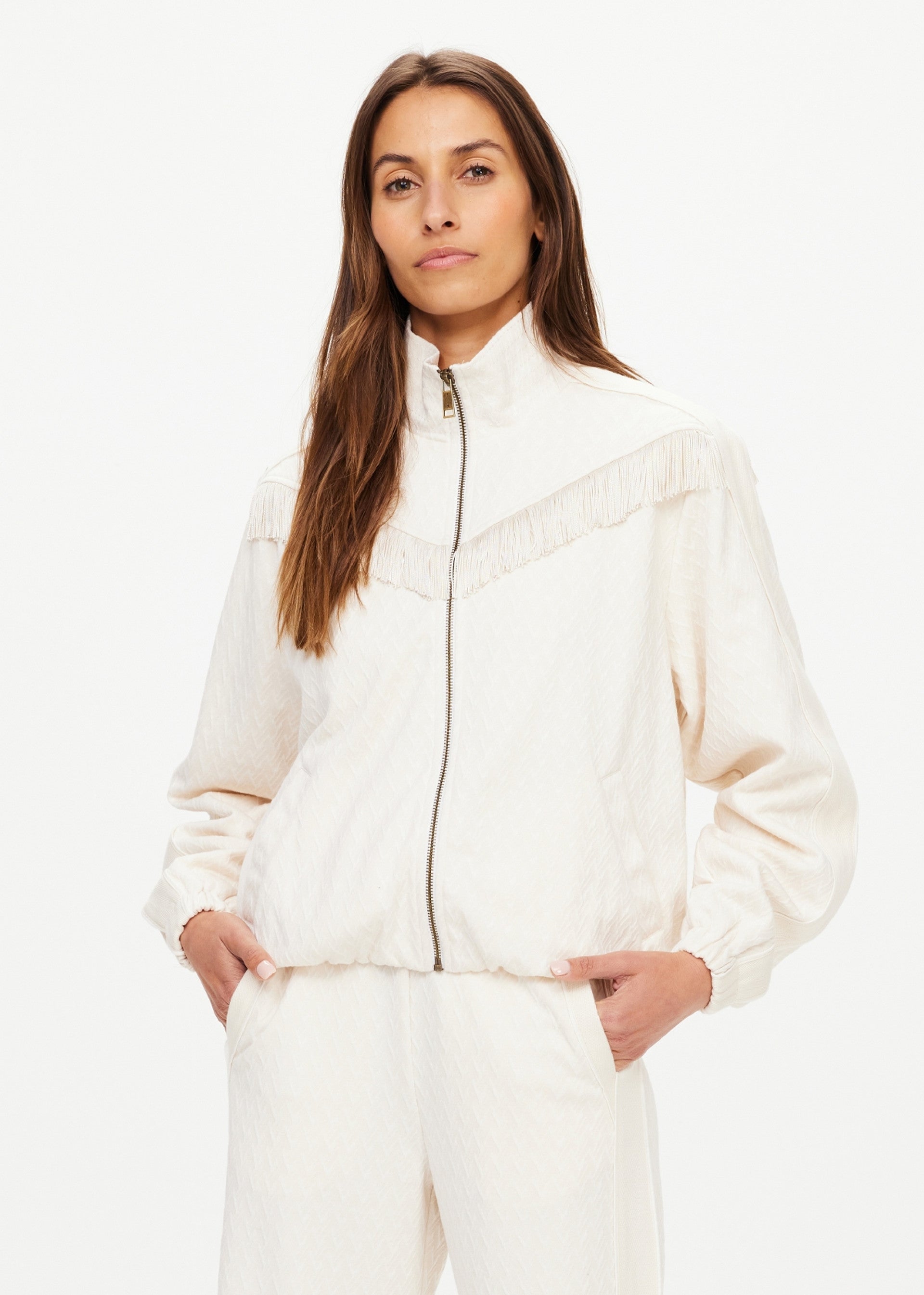 A woman with long brown hair wears The Upside’s BOW N ARROW FRINGE ZIP JACKET, a light-toned jacket with geometric design and fringe, paired with matching pants, standing hands-in-pockets against a plain white background.
