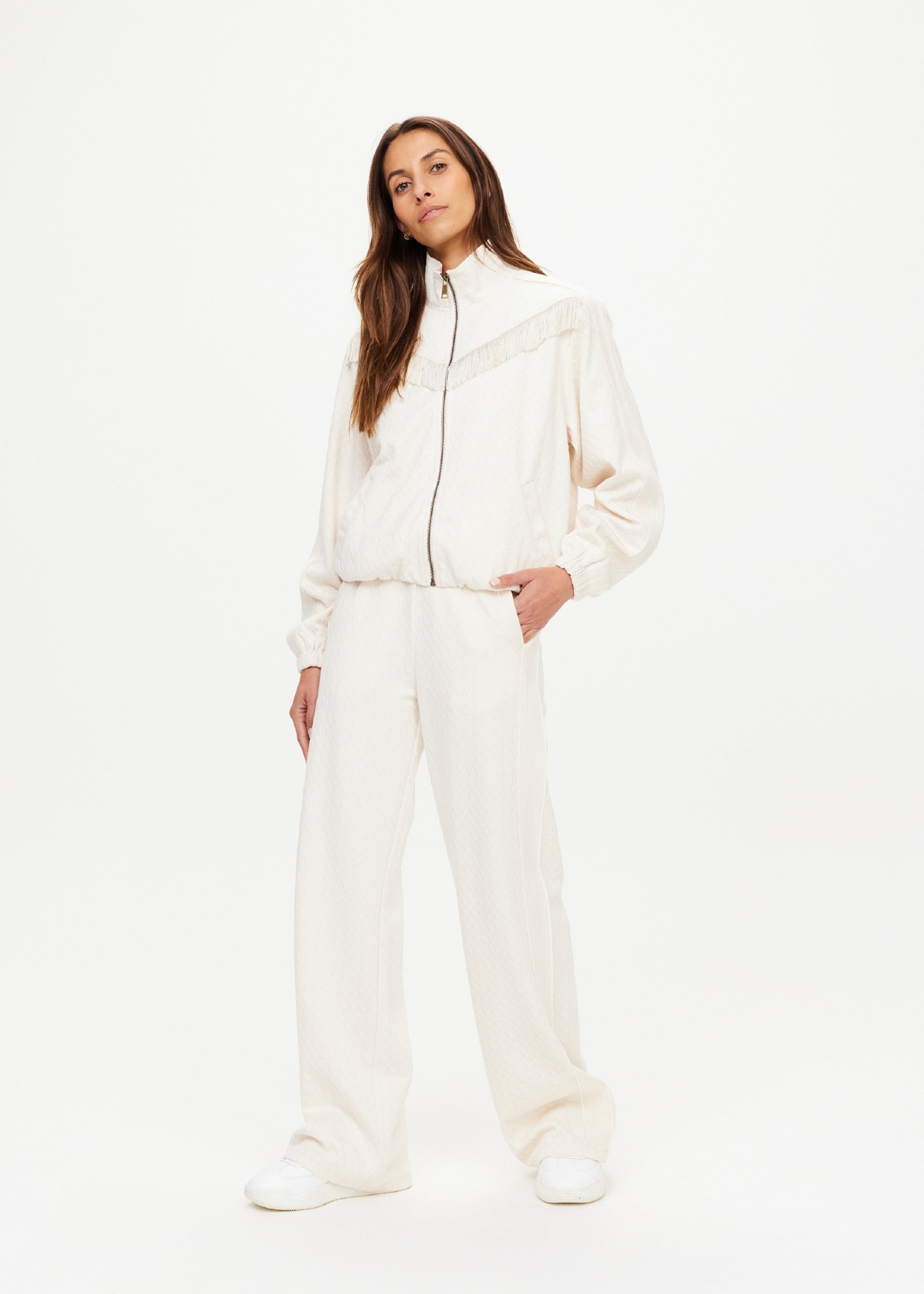 A woman with long brown hair stands confidently against a white background, wearing The Upside's BOW N ARROW FRINGE ZIP JACKET and matching wide-leg pants with white sneakers. Her hands are in her pockets as she looks at the camera.