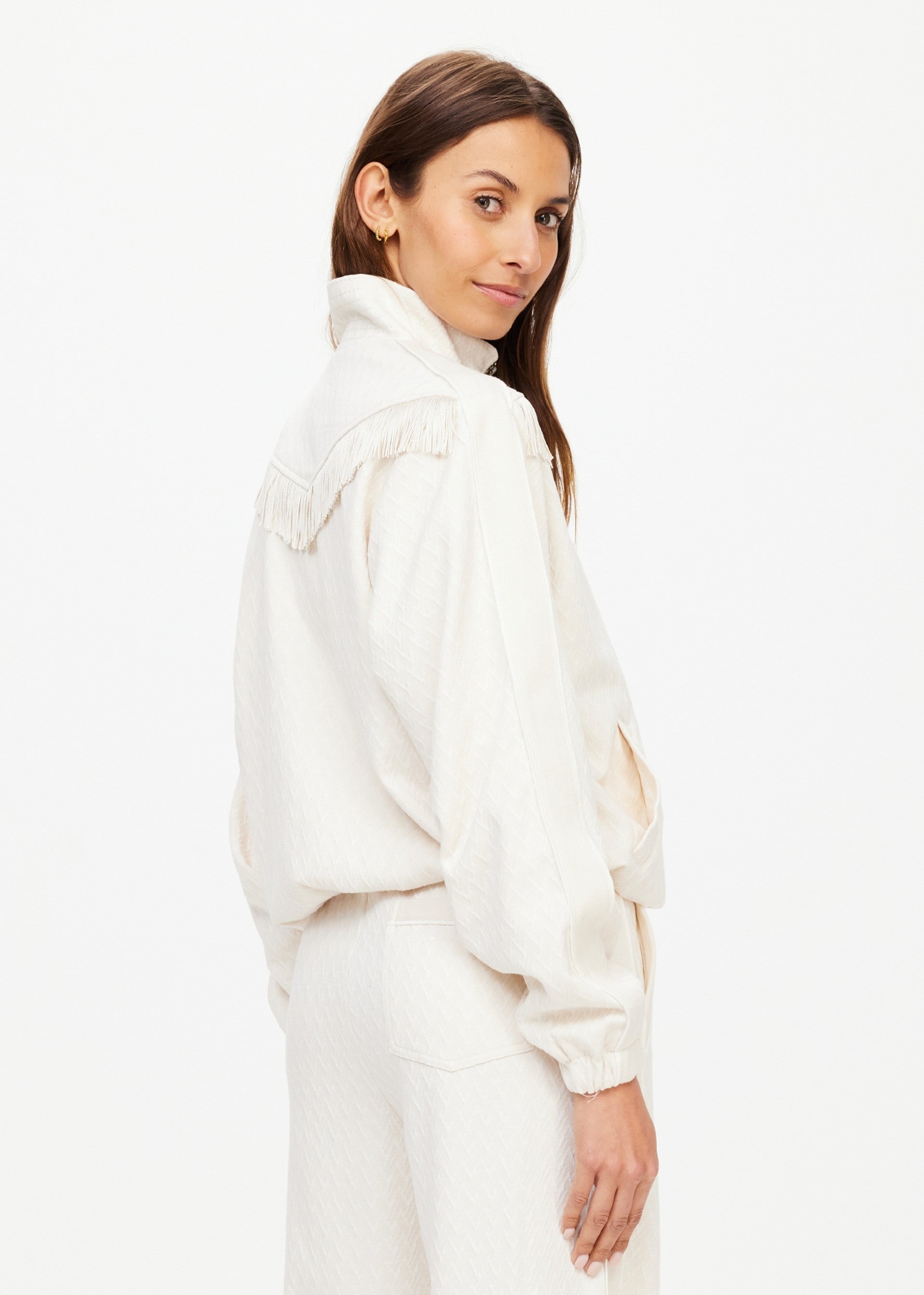 A woman with long brown hair wears The Upside's BOW N ARROW FRINGE ZIP JACKET, an off-white piece with a high collar and subtle geometric design, standing sideways and looking over her shoulder against a white background.