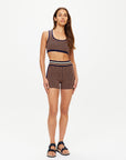 A woman with long brown hair and a neutral expression stands against a white background wearing The Upside's CROSSROAD 3IN KNIT SHORT, a patterned organic cotton short with an elasticated waistband, black sandals, and a matching crop top.