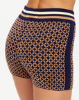 A close-up shows a person wearing The Upside’s CROSSROAD 3IN KNIT SHORT, featuring a high-waisted, elasticated waistband and a geometric navy blue and tan jacquard pattern.