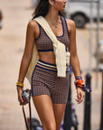 A woman wears The Upside's CROSSROAD 3IN KNIT SHORT with a matching patterned jacquard sports bra and elastic waistband, carrying a camera outdoors. She layers necklaces, bracelets, and colorful scarves, with a cream sweater draped over her shoulders.