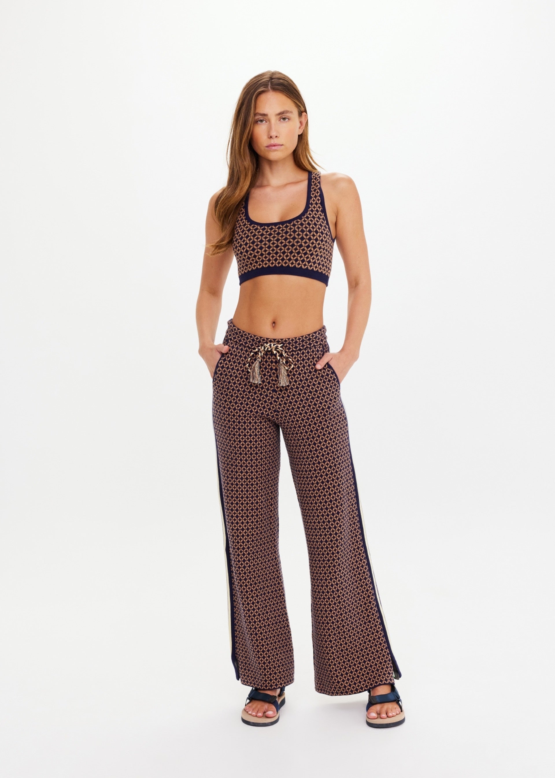 A woman with long hair stands facing forward in The Upside CROSSROAD JUNIPER KNIT PANT and a geometric sports bra, hands in pockets, wearing sandals against a plain white background.