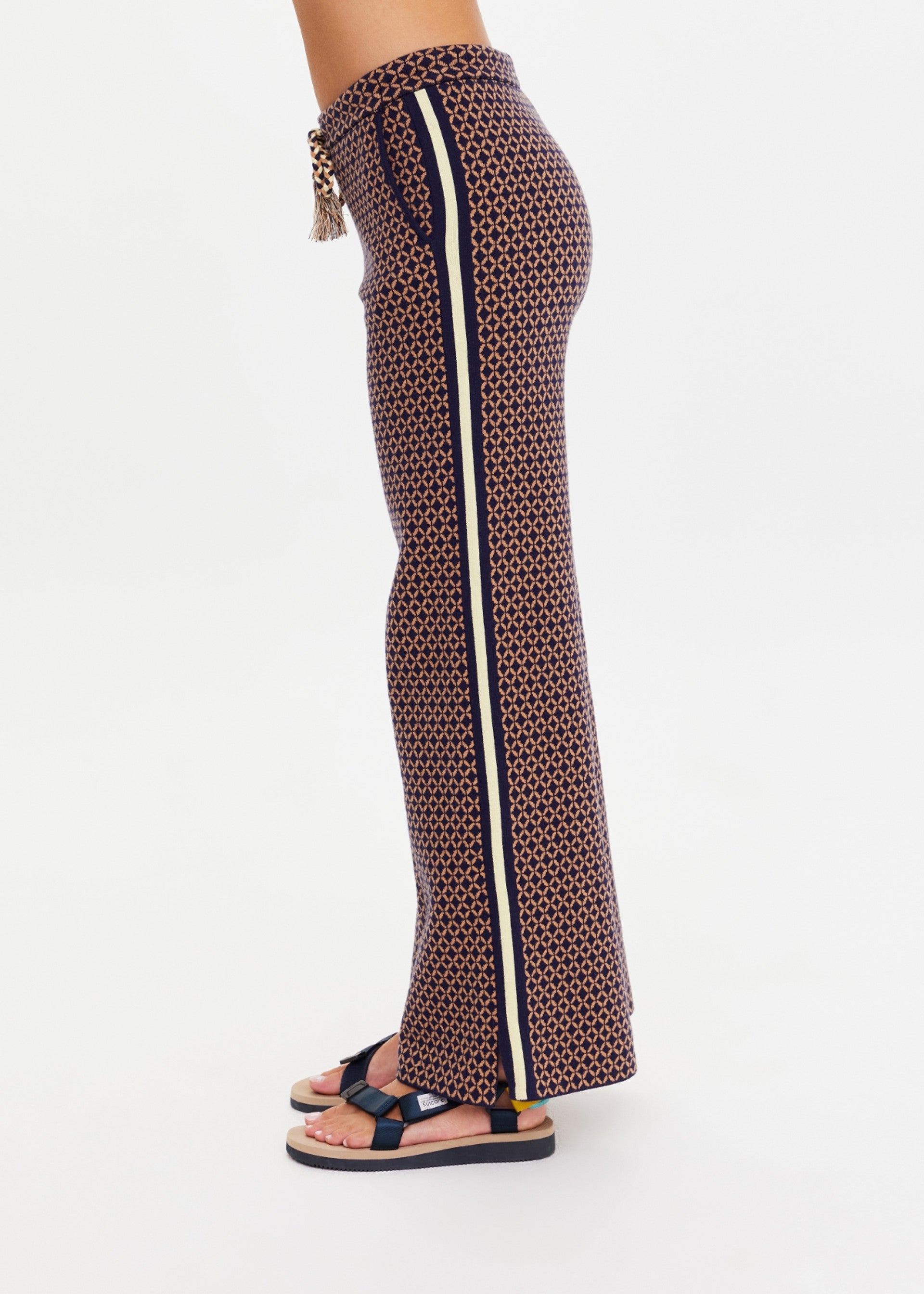 A person stands in profile against a white background wearing The Upside’s CROSSROAD JUNIPER KNIT PANT—wide-leg knit pants with a geometric design and vertical side stripe—paired with black platform sandals.