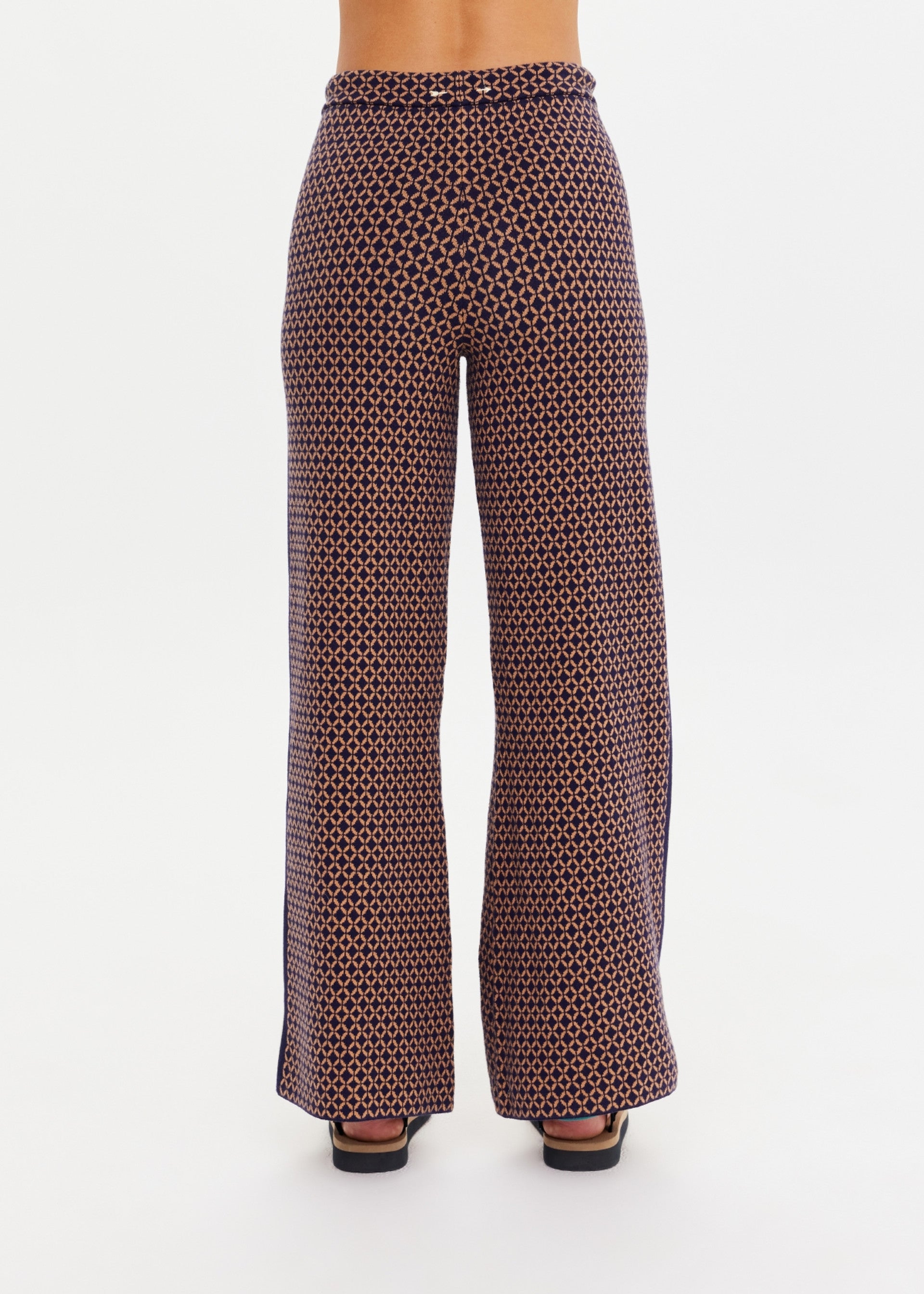 A model is shown from the back wearing The Upside CROSSROAD JUNIPER KNIT PANT—high-waisted, wide-leg pants featuring a black and brown geometric circular pattern—against a white background.