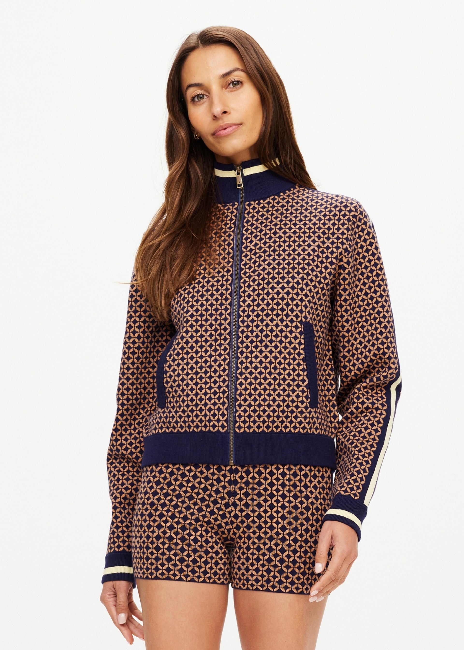 A woman with long brown hair wears The Upside CROSSROAD MARGOT KNIT JACKET, featuring a geometric design in brown and navy, paired with matching shorts, standing against a plain white background.