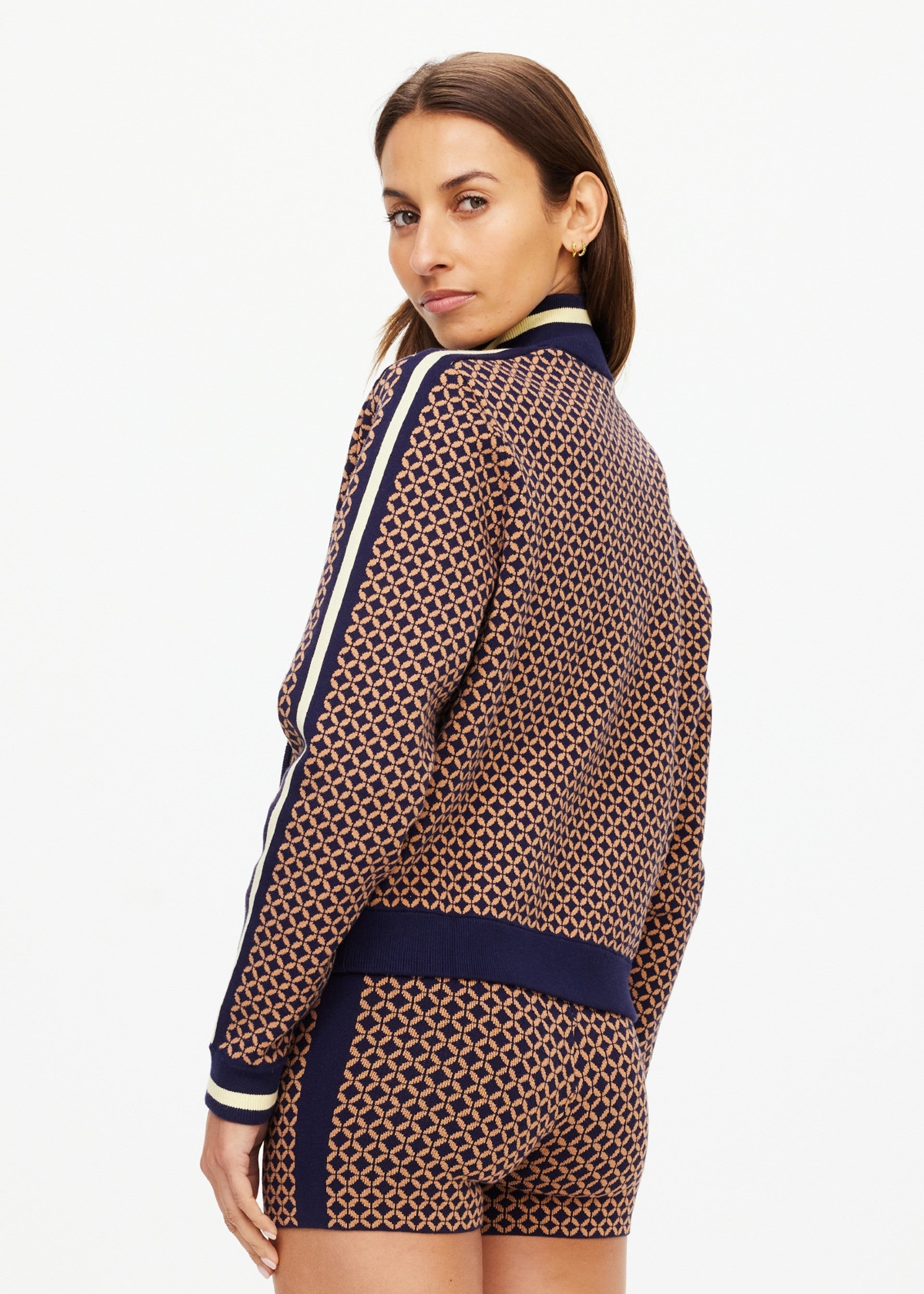 A woman looks over her shoulder, wearing The Upside’s CROSSROAD MARGOT KNIT JACKET and matching shorts in navy blue and beige geometric print with white stripes on the sleeves and shorts.