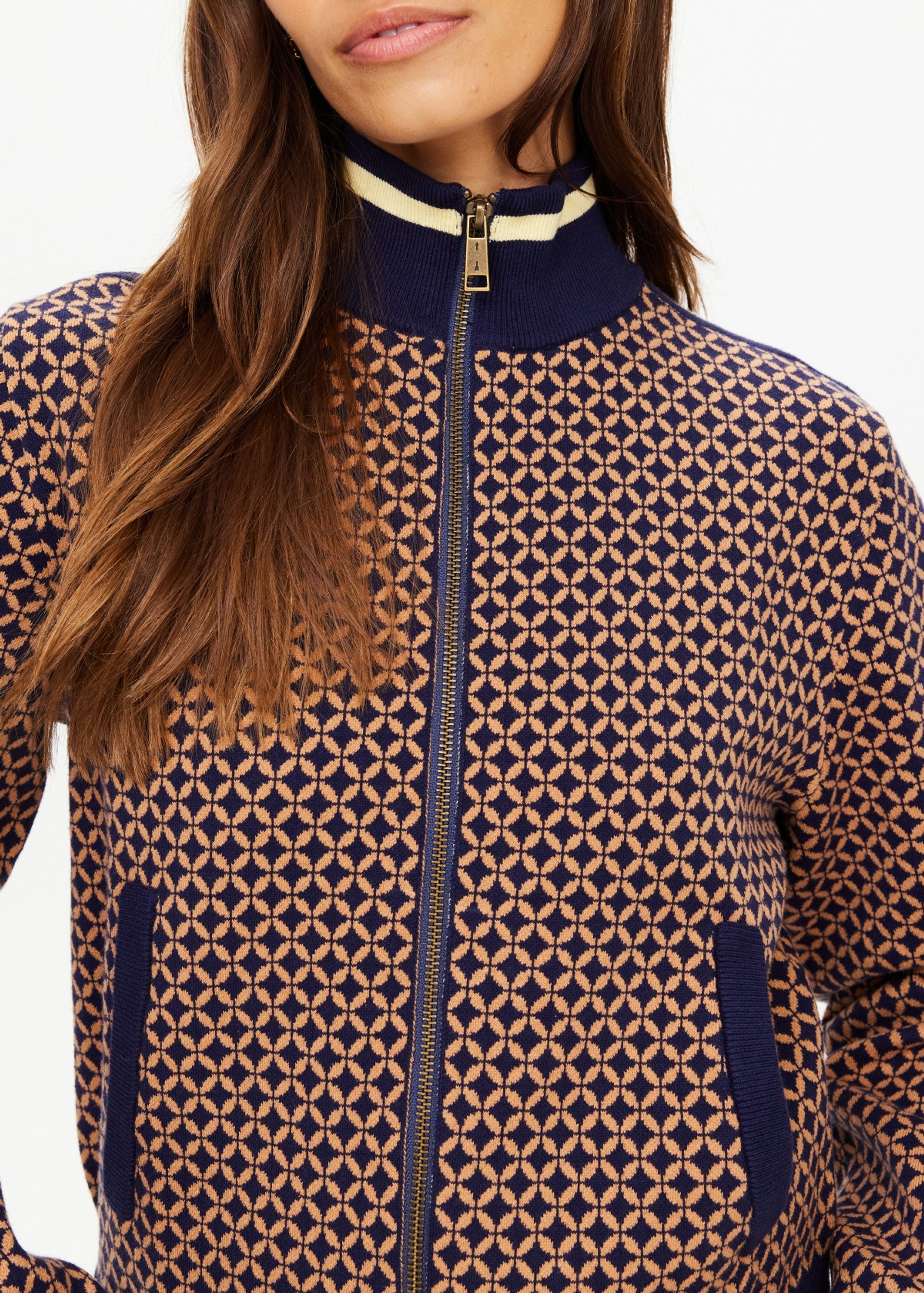 A woman models The Upside’s CROSSROAD MARGOT KNIT JACKET, a lightweight organic cotton blend with navy and tan geometric print, navy collar with white stripe, and navy-trimmed pockets. Photo is cropped above her nose and below her waist.