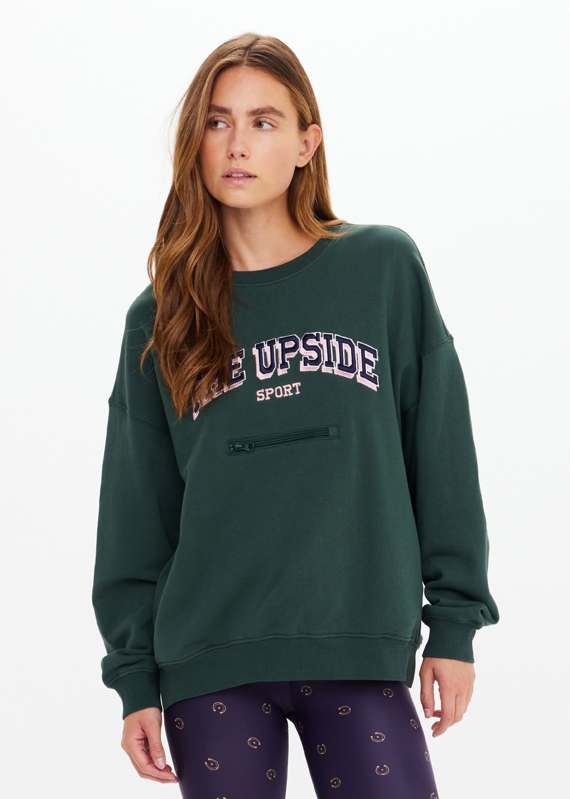 A woman with long brown hair wears The Upside’s ALUMNI COOLUM CREW organic cotton sweatshirt in dark green and patterned black leggings, standing against a plain white background.