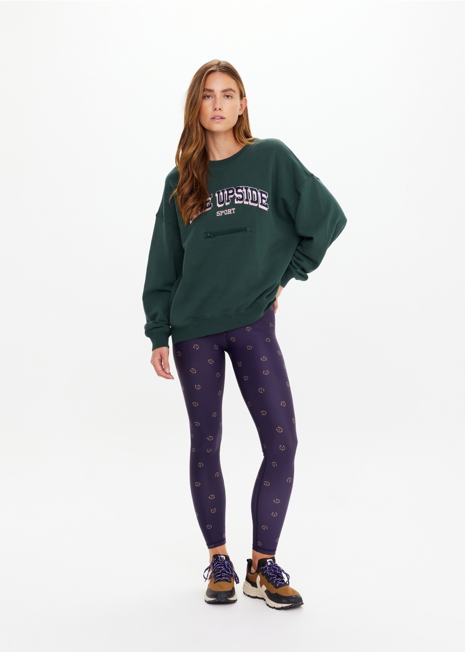 A woman with long, loose hair and a neutral expression stands against a white background wearing The Upside's ALUMNI COOLUM CREW sweatshirt in dark green, navy leggings with a small pattern, and brown sneakers with white soles.
